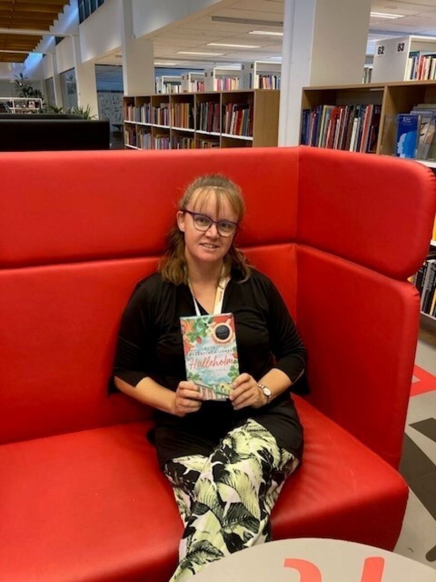 Lene sidder i en rød sofa med bogen "Halleholm - Lovisas valg" af Ruth Kvarnström-Jones
